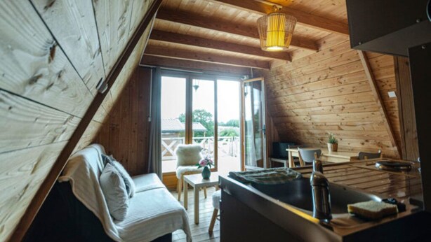 Een gezellig slaapkamer op zolder met een houten schuine dak, een bed, een klein tafeltje met een stoel en een raam met uitzicht op een tuin.