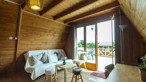 Een gezellig houten blokhutinterieur met een bank, tafel, stoelen en een grote schuifdeur naar een buitengebied.