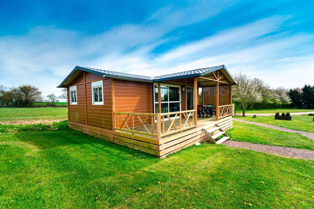 Een houten chalet met veranda en een klein terras, omgeven door een grasveld en bomen.
