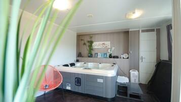 A modern bathroom with a large bathtub, a small sink, and a shower area. There are plants and decorative items on the wall, and a towel rack is visible.