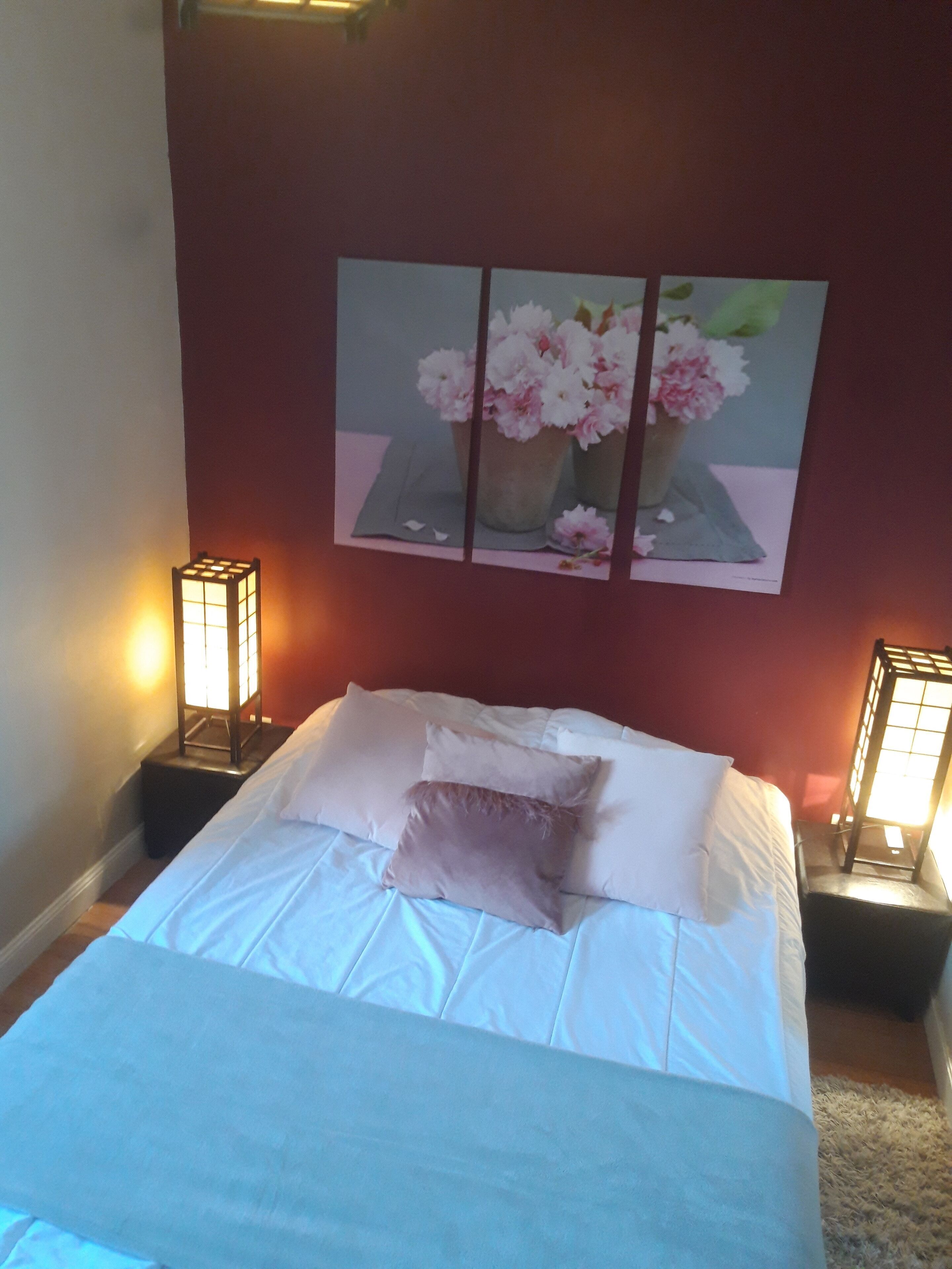 A neatly made bed with white linens and a single decorative pillow, flanked by two bedside tables with illuminated lamps. Above the bed, a triptych painting of flowers is displayed on the wall.