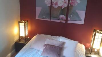 A neatly made bed with white linens and a single decorative pillow, flanked by two bedside tables with illuminated lamps. Above the bed, a triptych painting of flowers is displayed on the wall.