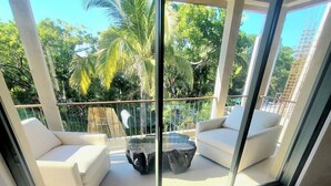 A balcony with white cushioned seating and a glass table, overlooking a lush green landscape with palm trees.