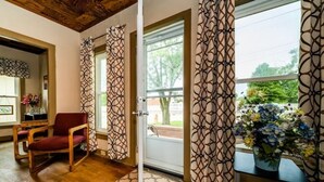 Une pièce avec un plancher en bois, une chaise rouge, une porte ornée de rideaux à motifs et une fenêtre donnant sur un paysage verdoyant.