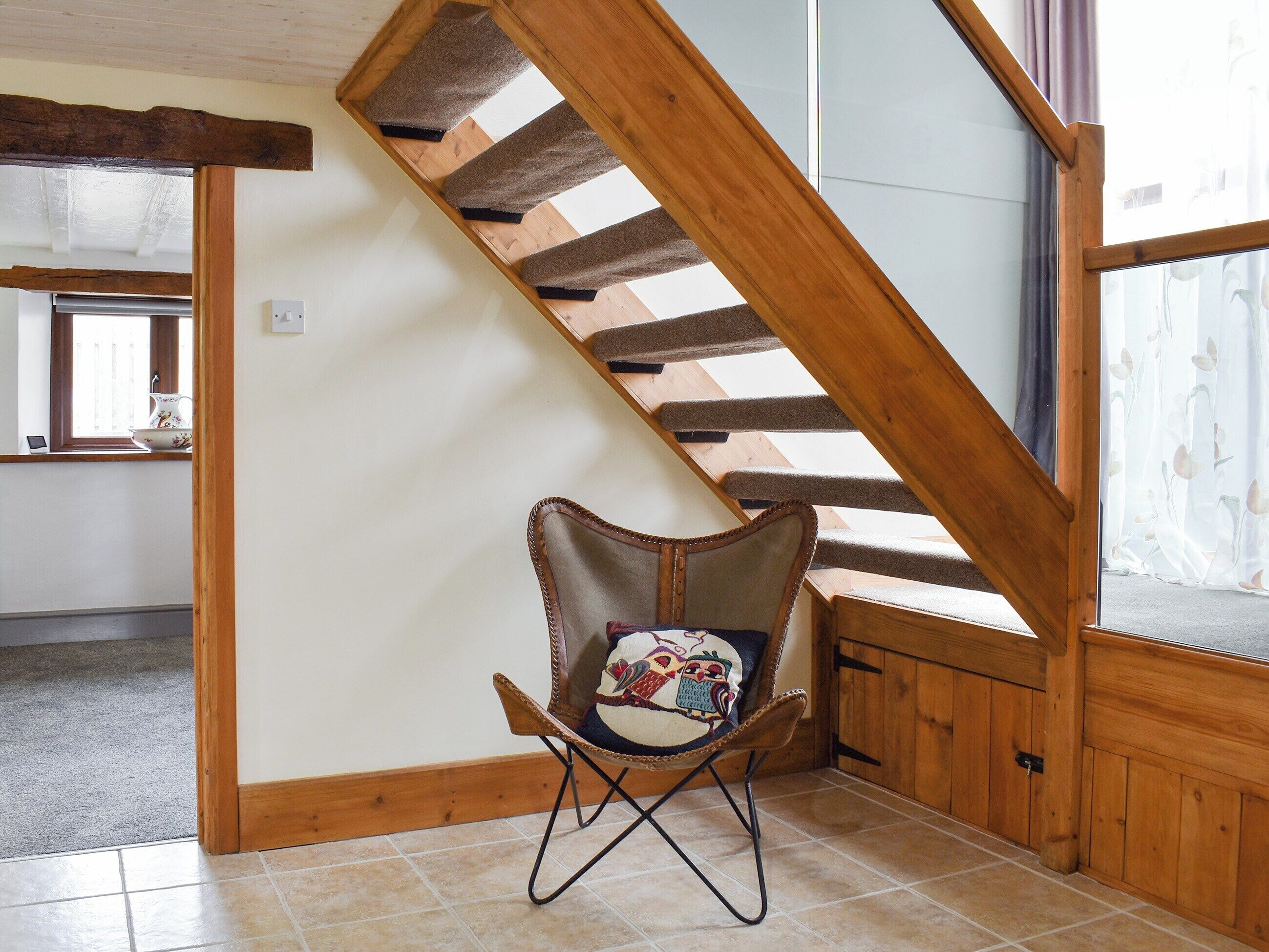 Una escalera de madera con una butaca acolchada en una habitación con suelo de baldosas y una ventana con cortinas florales.