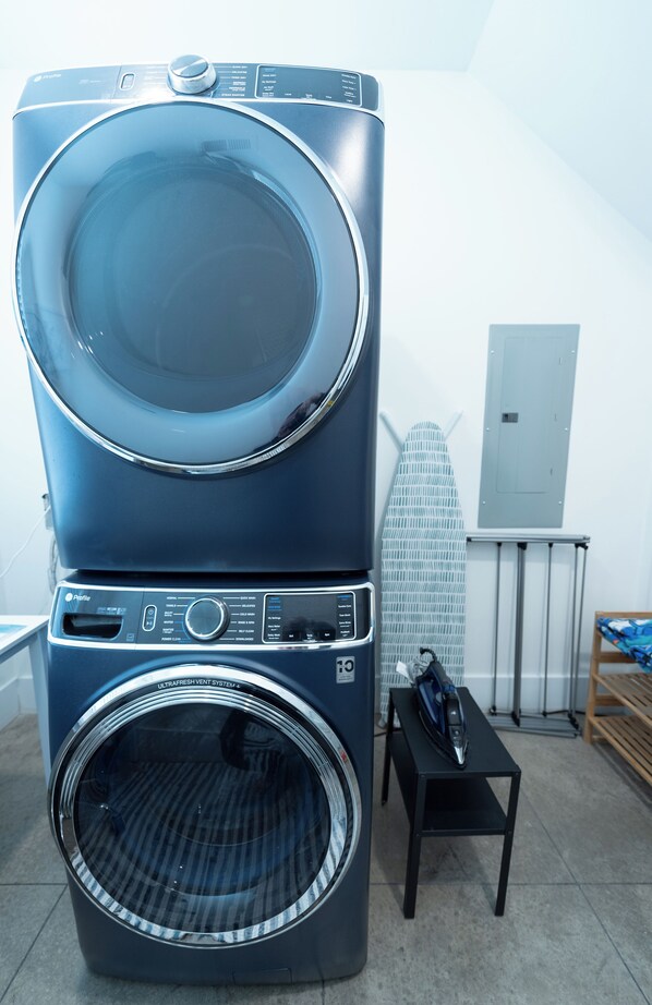 A stacked washer and dryer unit with a control panel, located in a room with a tiled floor, ironing board, and a small table with an iron.