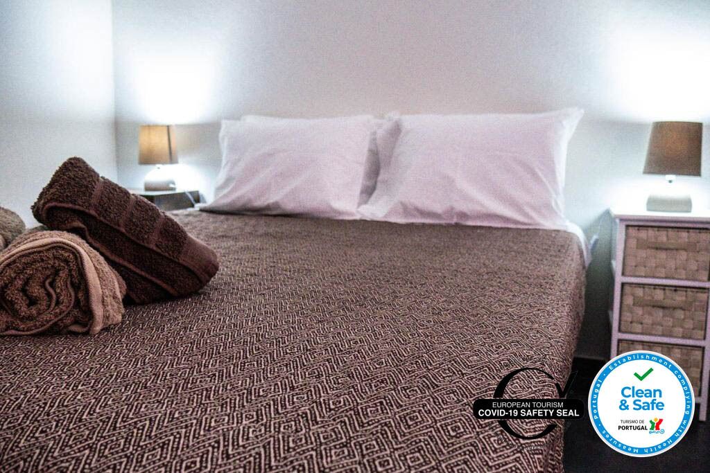 A hotel room with a patterned bedspread, two white pillows, a rolled-up towel, and a nightstand with a lamp. The room has a 'Clean & Safe' COVID-19 safety seal.