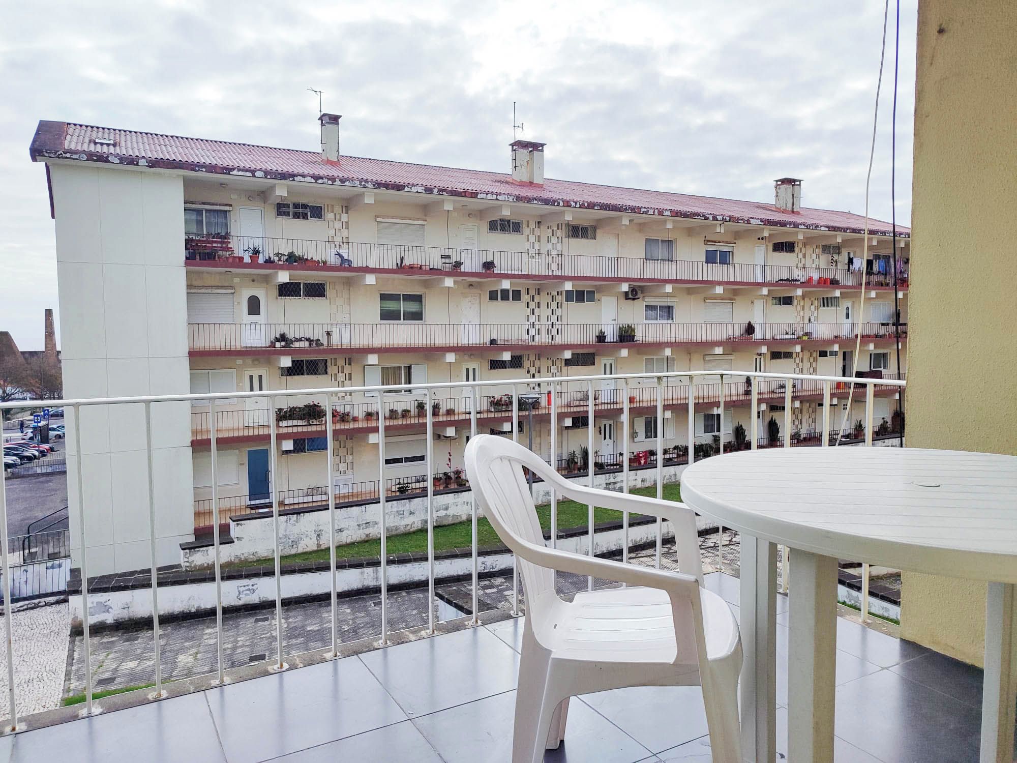 Ein Balkon mit weißem Plastikstuhl und Tisch, der auf ein mehrstöckiges Gebäude mit Balkonen blickt.