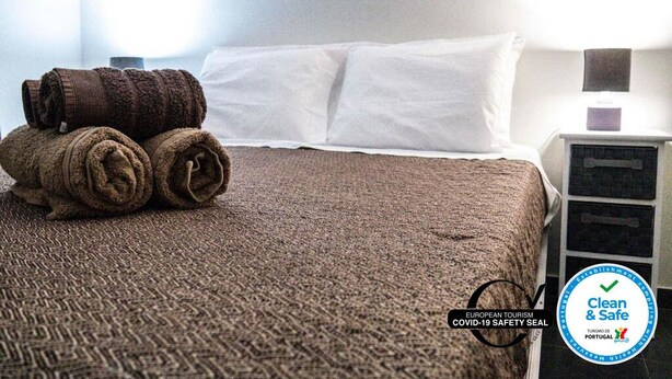 A hotel room with a neatly made bed, a rolled-up towel, and a bedside table with a lamp. The room has a clean and safe environment as indicated by the signage. - Bernardo's Place (Ponta Delgada)