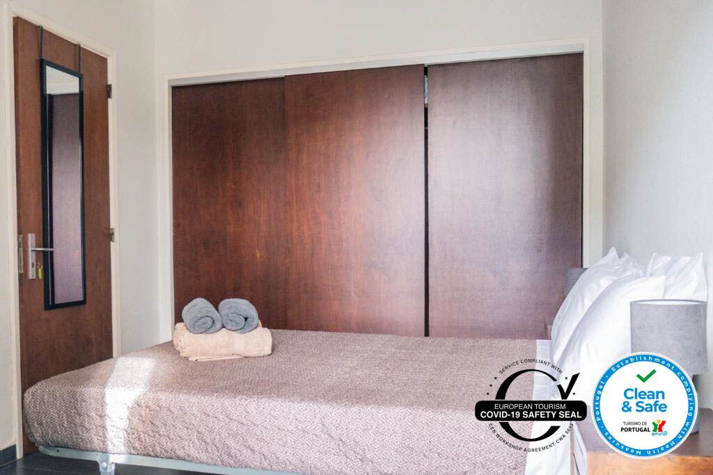 Une chambre d’hôtel avec une grande armoire en bois, un lit recouvert d’une courtepointe à motifs et une pile de serviettes enroulées posée sur le lit. On aperçoit un oreiller blanc sur le lit et un autre oreiller blanc sur le côté droit. La chambre est propre et offre un environnement sûr, comme l’indiquent les panneaux d’information.