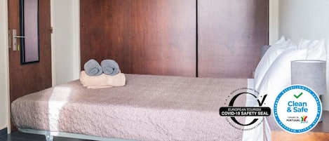 Une chambre d’hôtel avec une grande armoire en bois, un lit recouvert d’une courtepointe à motifs et une pile de serviettes enroulées posée sur le lit. On aperçoit un oreiller blanc sur le lit et un autre oreiller blanc sur le côté droit. La chambre est propre et offre un environnement sûr, comme l’indiquent les panneaux d’information.