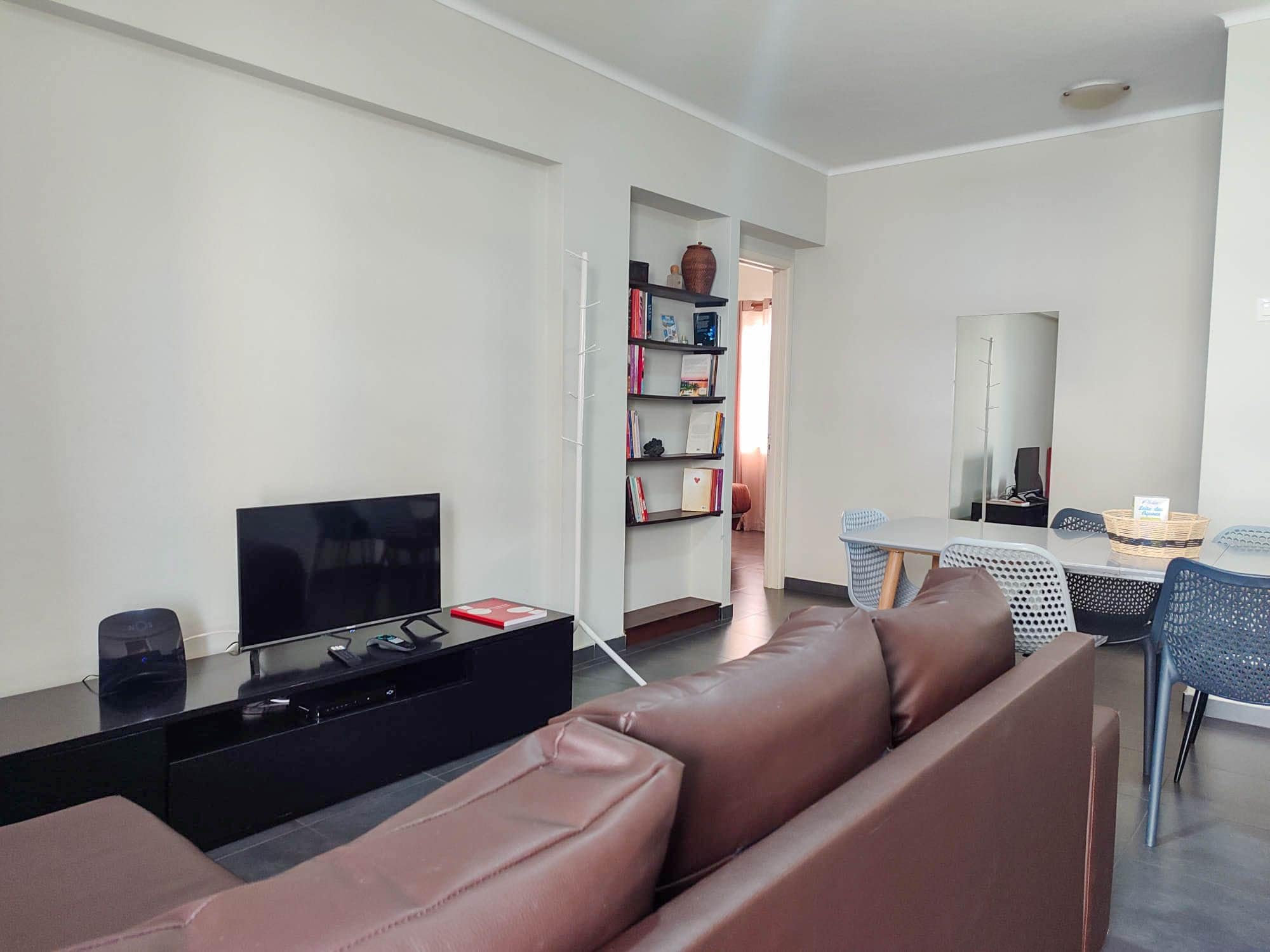 Una sala de estar moderna con un televisor de pantalla plana, un mueble de entretenimiento negro, un sofá de cuero marrón, una zona de comedor con mesa y sillas, y una estantería con diversos objetos.