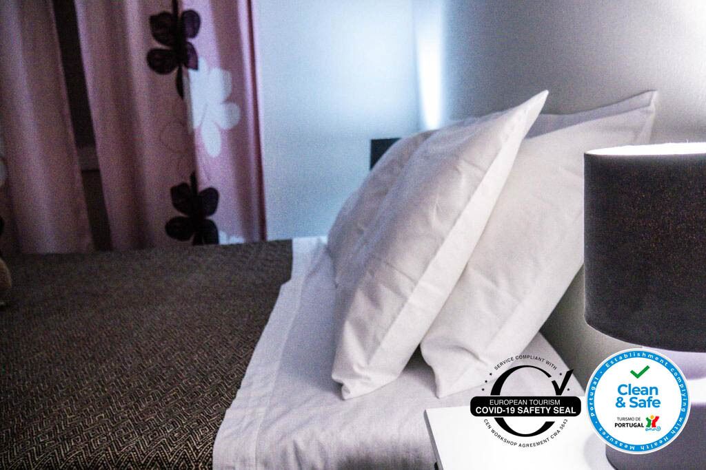 Habitación de hotel con cama, almohadas, mesita de noche y lámpara. Se observan logotipos que indican cumplimiento de medidas de seguridad por COVID-19 y una certificación 'Clean & Safe'.