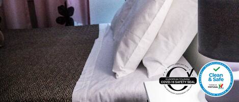 Habitación de hotel con cama, almohadas, mesita de noche y lámpara. Se observan logotipos que indican cumplimiento de medidas de seguridad por COVID-19 y una certificación 'Clean & Safe'.