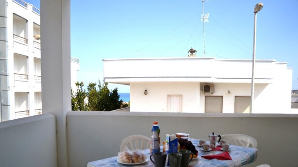 A balcony with a table set for two, white plastic chairs, and a view of a building and the sea. - Charming Holiday Home in San Foca, Italy - Perfect for Families (Melendugno)