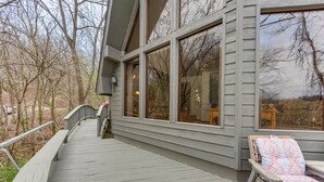 A wooden deck with a railing, a chair with a patterned cushion, and a large window overlooking a wooded area. - Riverfront Baxter Springs Home w/ Deck & Grill! (Baxter Springs)