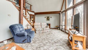 A spacious living room with a high ceiling, a large window, a blue armchair, a patterned sofa, and a wooden side table with a lamp. - Riverfront Baxter Springs Home w/ Deck & Grill! (Baxter Springs)
