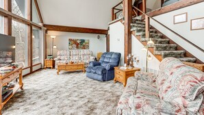 A spacious living room with a high ceiling, a large window, a blue armchair, a patterned sofa, and a staircase with wooden railings. - Riverfront Baxter Springs Home w/ Deck & Grill! (Baxter Springs)