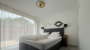 A bedroom with a bed, a nightstand, a wall-mounted lamp, and a window with curtains. - Gite Les Mirettes Entre Terre et mer Avec Piscine, Salle de Jeux (Pornic)