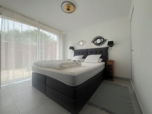 A bedroom with a bed, a nightstand, a wall-mounted lamp, and a window with curtains.