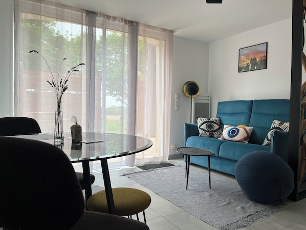 A living room with a round glass table, a blue sofa with patterned cushions, a round floor lamp, and a painting on the wall.