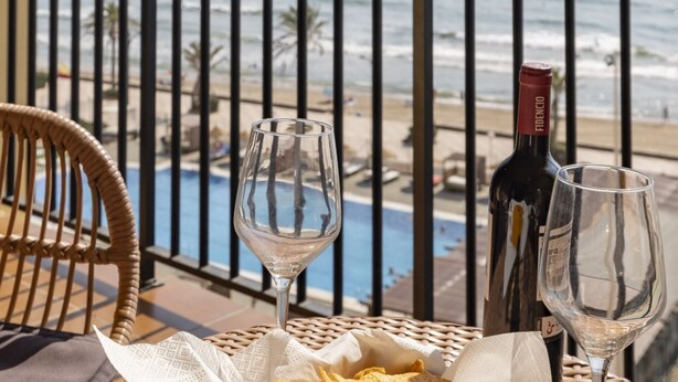 A balcony with a wicker chair, two wine glasses, a bottle of wine, and a pool view. - A (VA068) TH Port Saplaya con vistas al Mar. (Alboraya / Alboraia)