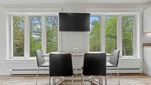 Un área de comedor con una mesa y sillas, un televisor de pantalla plana montado sobre la mesa, y ventanas grandes con vistas a árboles.