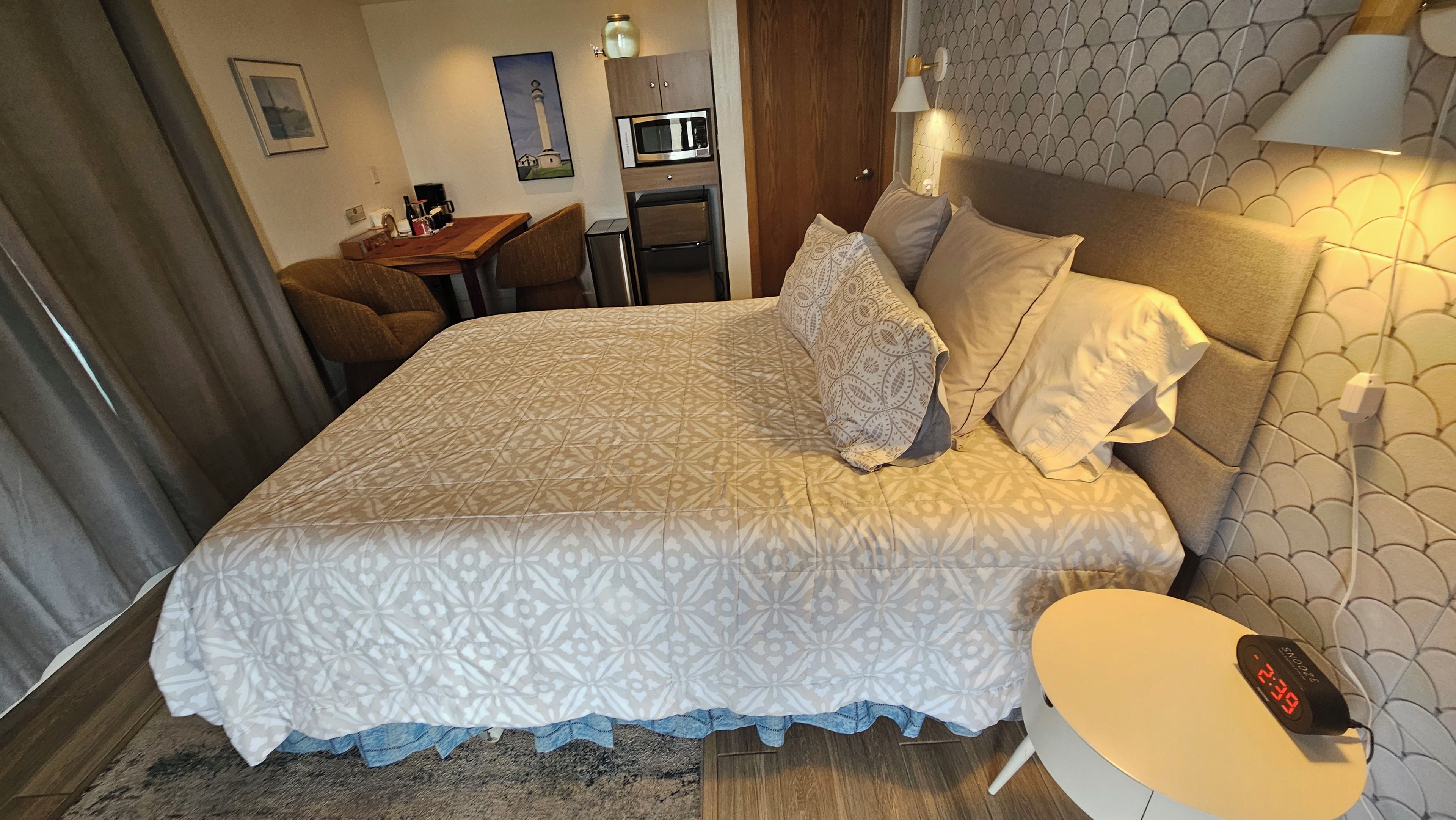 A hotel room with a bed, bedside table, lamp, and a desk with a chair.