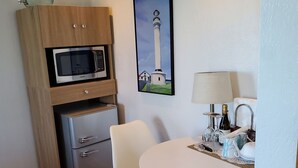 A compact kitchen with a microwave, refrigerator, and a small cabinet. There's a dining table with chairs and a painting of a lighthouse. - Point Arena Lighthouse - Keeper's Room (Point Arena)
