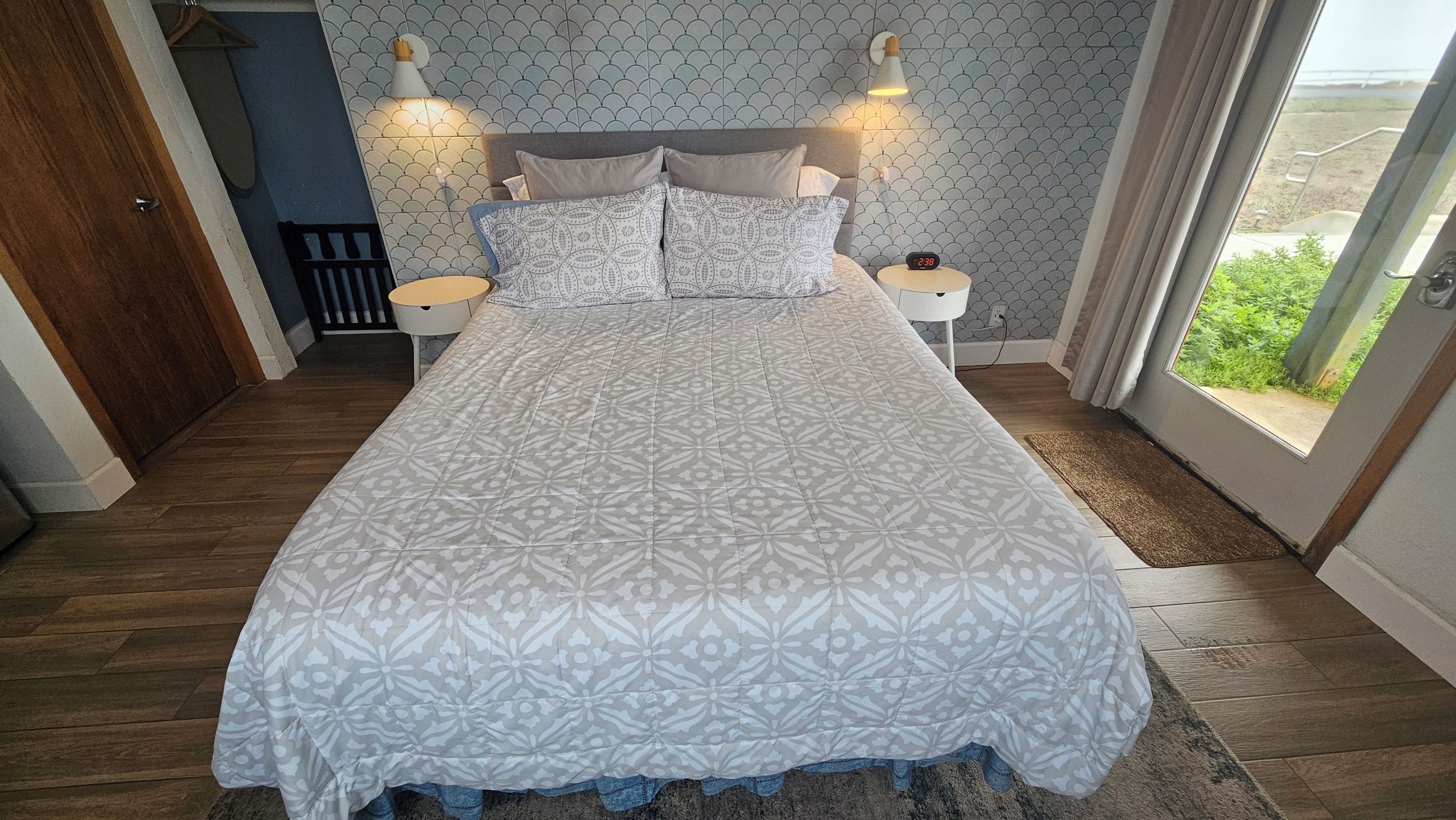 A bedroom with a bed, two bedside tables, a nightstand, a wall clock, and a window with a view.