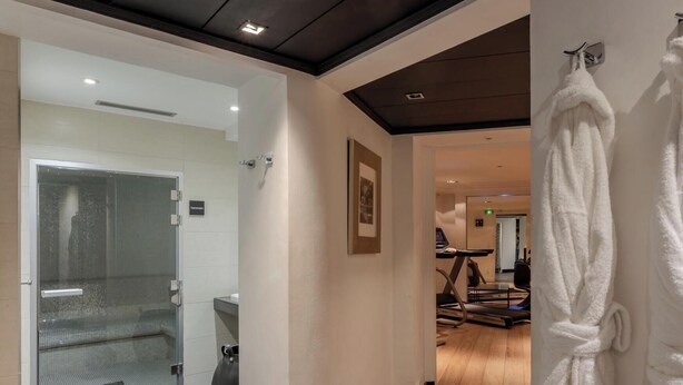 A hotel corridor with a glass-enclosed shower area, a treadmill in the background, and a white robe hanging on the wall. - Hotiday Room Collection - Paris Arc de Triomphe (Paris)