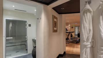 A hotel hallway with a glass door leading to a bathroom, a treadmill, and a rack of white robes.