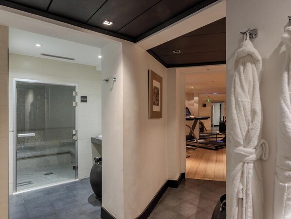 A hotel hallway with a glass door leading to a bathroom, a treadmill, and a rack of white robes.