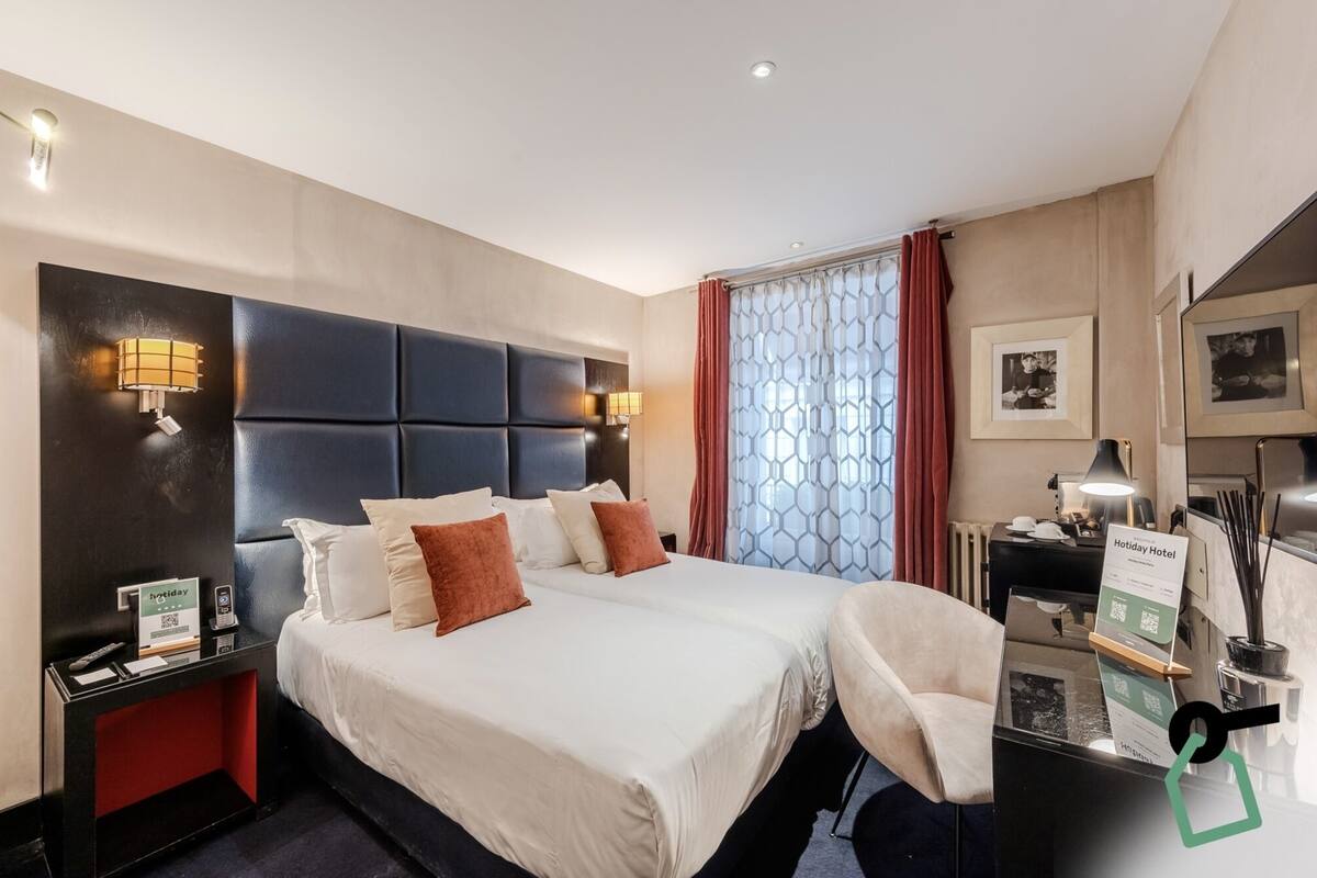 Ein Hotelzimmer mit einem großen Bett, zwei Nachttischen, einem Schreibtisch mit Stuhl, einer Minibar und einem Fenster mit Vorhängen.