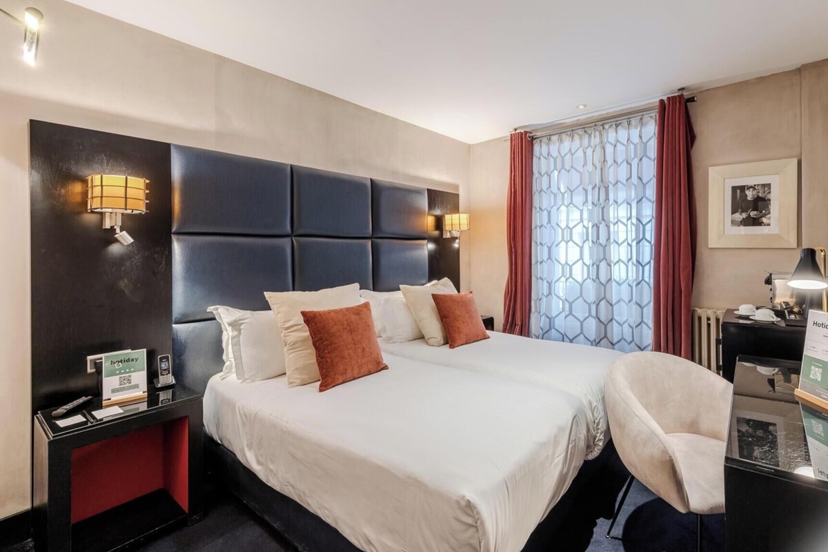 A hotel room with a large bed, two bedside tables, a desk with a chair, a minibar, and a window with curtains.