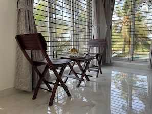 A room with a view of palm trees through barred windows, a wooden table with a banana and a cup, and two wooden chairs. - Gintara Homestay by evaddo stays (Udupi)