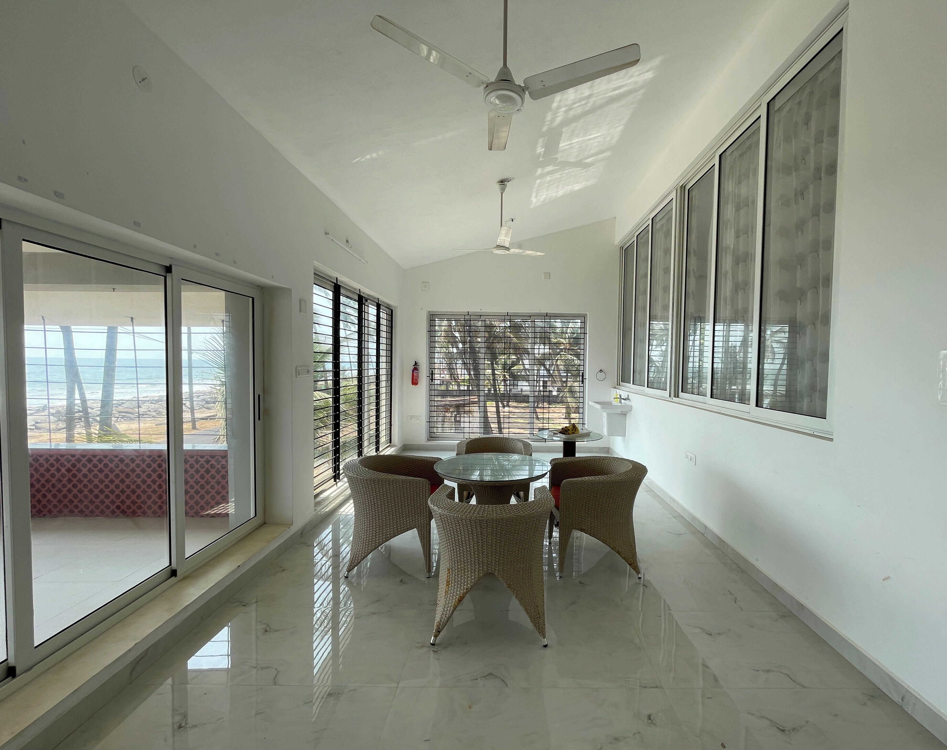 Amplia habitación con una gran mesa de comedor rodeada de sillas de mimbre, un ventilador de techo y ventanas amplias que ofrecen vistas al exterior.