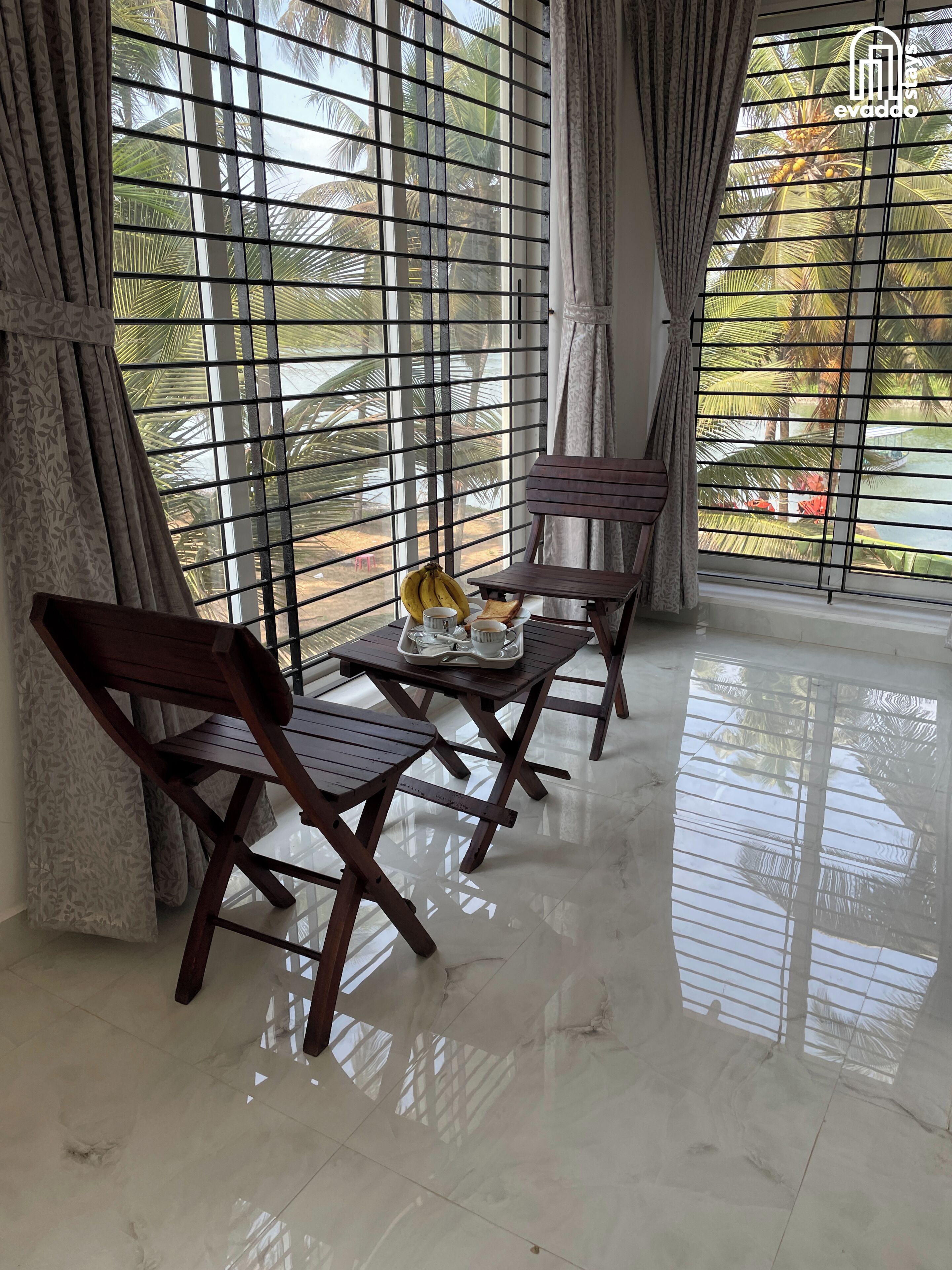 A room with a view of palm trees through barred windows, a table with a tray of bananas, and wooden chairs.