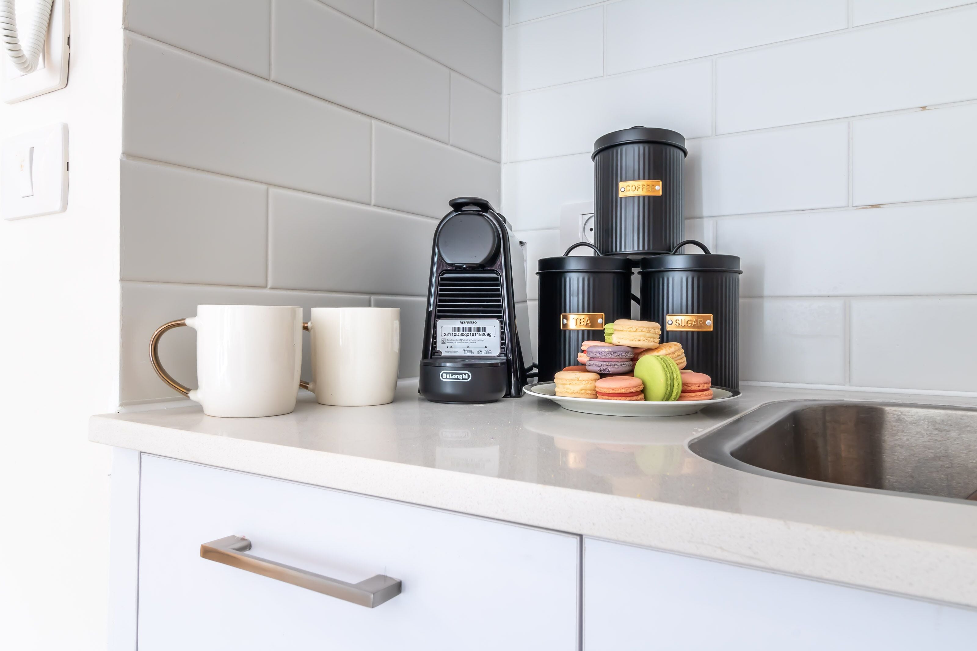 A kitchen counter with a coffee maker, two thermos flasks, and a plate of macarons.