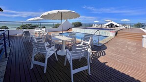 Ein Poolbereich mit weißen Plastikstühlen und Sonnenschirmen, bewölkter Himmel und Blick auf das Meer.