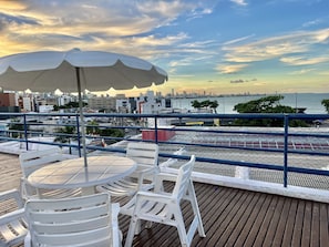 A rooftop terrace with a table and chairs, a large umbrella, and a view of a city skyline and water. - Manos Hotel Flat 118 (João Pessoa)