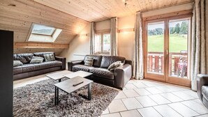 A cozy living room with a wooden ceiling, two leather sofas, a coffee table, and a large sliding glass door leading to an outdoor area. - Wifi, 200m from ski slopes, 4th floor, balcony, tv, ski locker, 105m², Les Gets (Les Gets)