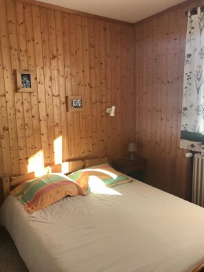 Ein Zimmer mit Holzvertäfelung, einem Bett, einem Nachttisch und einem Fenster mit floralem Vorhang.