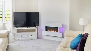 Uma sala de estar com uma televisão de tela plana sobre um suporte branco, uma lareira branca com fogo aceso e um sofá bege com almofadas coloridas.