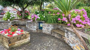 Jardim com paredes de pedra, um canteiro de madeira cheio de flores coloridas, uma placa de aviso e um caminho de pedras.