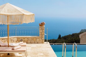 Zona junto a la piscina con un muro de piedra, una sombrilla tejida, sillas de descanso y vista al mar.