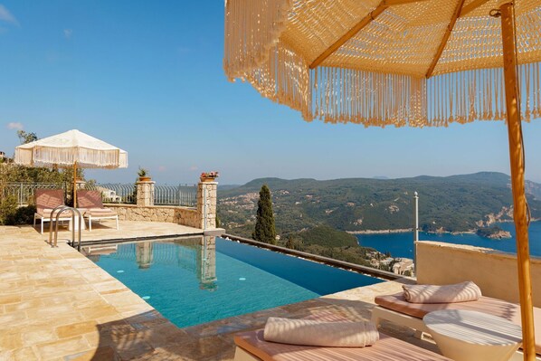 Una piscina infinita con vistas despejadas al mar y las colinas, suelo de piedra, sillones de descanso y una pérgola con toldo.