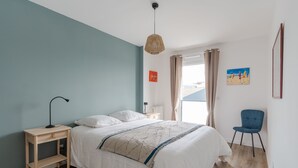 A bedroom with a bed, bedside table, lamp, chair, and artwork on the wall. - Madreperla - Appt à moins d'un km de la plage (Courseulles-sur-Mer)