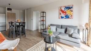 A modern living room with a grey sofa, a dining table with chairs, a black coffee table with a vase, and a colorful abstract painting on the wall. - Madreperla - Appt à moins d'un km de la plage (Courseulles-sur-Mer)