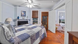 Une chambre à coucher avec un grand lit, un ventilateur de plafond, une cheminée et une porte donnant sur une autre pièce.